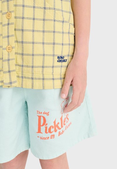 Enfant portant une chemise à carreaux jaunes boutonnée et un short bleu clair avec un motif de chien rouge « Pickles » sur la jambe gauche, main reposant sur le côté.