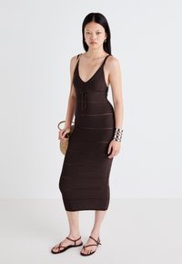 Ruda megzta midi suknelė su V iškirpte, reguliuojamu raišteliu talijoje ir horizontaliomis juostelėmis. Modelis dėvi plokščius sandalus ir laiko nertą krepšį.