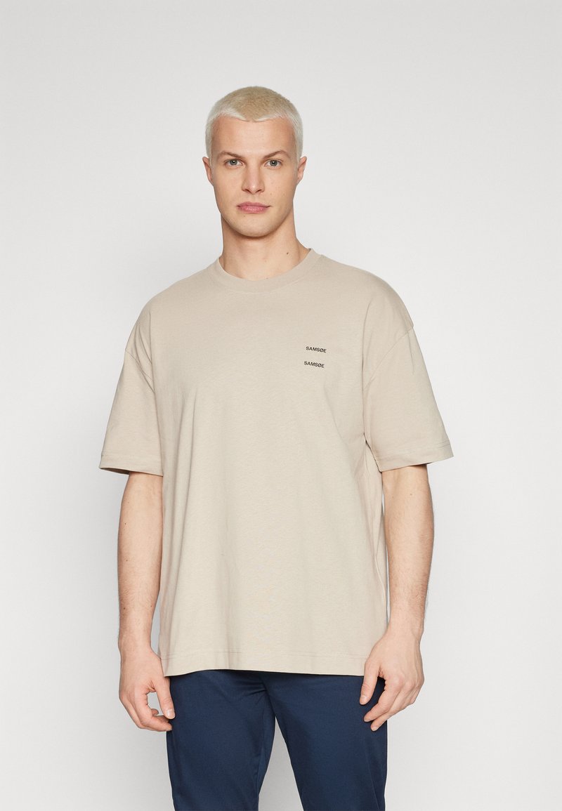 Beige kortermet t-skjorte laget av myk stoff, med en liten trykt logo i mørk tekst på brystet. Enkel, avslappet passform.