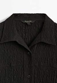 Chemise noire texturée avec col, deux boutons et détails de coutures horizontales, présentant l'étiquette de la marque "Massimo Dutti" au niveau de l'encolure.