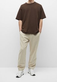 Person wearing a loose brown short-sleeve shirt, beige pants, and white sneakers, standing with hands in pockets against a plain background.