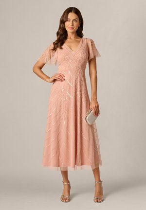 Kvinde i en blush pink, blomsterbroderet midi kjole, holder en lille sølv clutch, iført sølv åbne pejs sko, og står mod en ensfarvet baggrund.