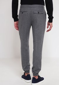 Man wearing grey tailored jogger pants with elastic cuffs and black buttoned back pockets, paired with navy sneakers and a black top.