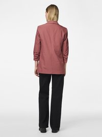 Femme aux cheveux blonds raides mi-longs, portant un blazer rose poudré et un pantalon noir à jambes larges, debout de dos sur un fond blanc.