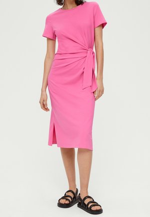Vrouw draagt een felroze jurk tot op de knie met een strikdetail in de taille en een zijsplit, gecombineerd met zwarte sandalen met open teen.