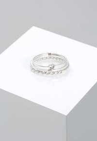 Tre anelli di argento intrecciati, con un design a spirale, uno dei quali presenta una piccola gemma incastonata. Superfici lisce, finitura lucida e aspetto brillante.