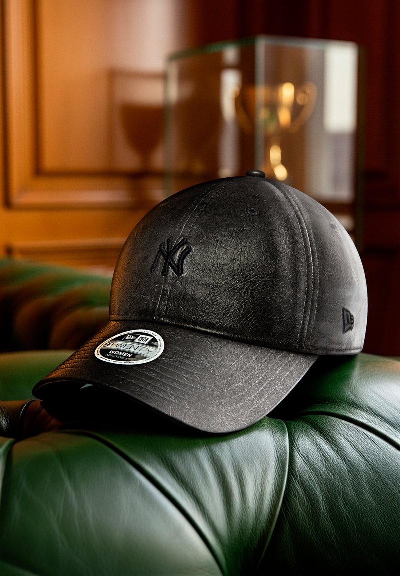 Zwarte leren New York Yankees honkbalpet met verstelbare sticker, rustend op een groene leren stoel in een warm getinte kamer.