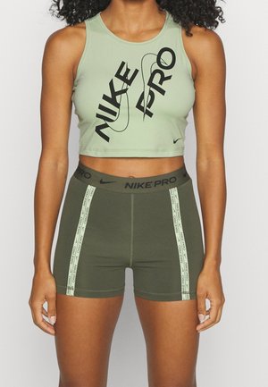 Kobieta nosząca jasnozielony krótki top Nike Pro z czarnym napisem i ciemnozielone spodenki Nike Pro z paskami z logo po bokach.