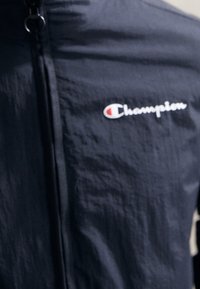 Černá nylonová bunda se zapínáním na zip a logem "Champion" v bílé a červené barvě na levé části hrudi. Hladká textura, lehký design.