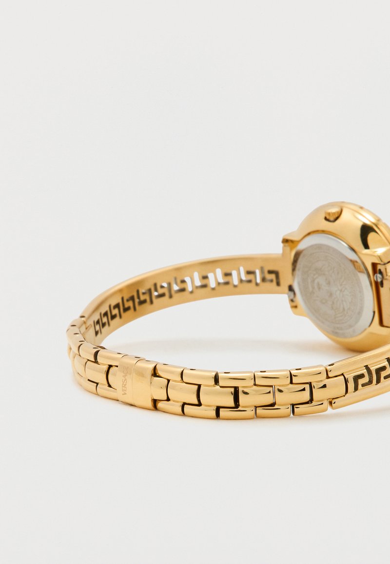 Gouden metalen polshorloge met een gegraveerde schakelband, ronde kast en transparante achterkant. Heeft zwarte accenten aan de binnenkant van de band.