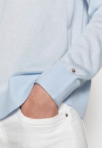Jasnoniebieski sweter dzianinowy z prążkowanymi mankietami, z małym akcentem logo. Umieszczony blisko białej kieszeni dla kontrastu i detalu.
