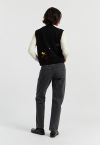 Zwarte gebreide vest met geborduurde ontwerpen van een gele vogel en een rood hart, gedragen over een crèmekleurig langetermijn shirt en gecombineerd met grijze spijkerbroek.