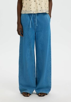 Personne portant un pantalon en jean bleu clair à jambes larges avec une taille à cordon et des sandales noires ouvertes aux orteils, debout les mains dans les poches.