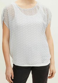 Femme portant un chemisier blanc à manches courtes avec un motif en vagues en demi-cercle noir, associé à un pantalon noir, visible du cou aux hanches.