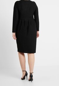 Czarna sukienka z długim rękawem o dopasowanej górze i plisowanej spódnicy, wykonana z teksturowanego materiału. Połączona z czarnymi sandałami na wysokim obcasie z paskami.