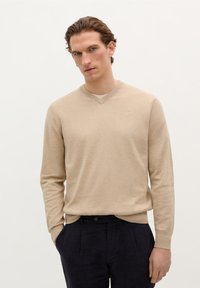 Beiger gestrickter Pullover mit V-Ausschnitt, langen Ärmeln und gerippten Bündchen. Über dunklen Hosen getragen, mit einem dezenten Logo auf der Brust.