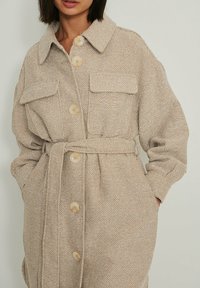 Beige textuurjas met een knoopsluiting aan de voorkant, grote kraag, twee borstzakken en een ceintuur in de taille. Zacht materiaal met een geweven patroon.