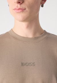 Barna, kerek nyakú póló, puha pamut anyagból, a mellkasán dombornyomott "BOSS" logóval. Simább textúra és testhezálló kialakítás.