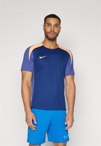 Moška športna majica Nike z modrim trupom, oranžnimi ramenskimi poudarki in kratkimi rokavi. V kombinaciji z modrimi kratkimi hlačami. Gladka tekstura materiala.