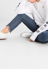 Sweat à capuche blanc avec logo, détails noirs sur les manches ; associé à un jean bleu déchiré et des baskets blanches avec une semelle texturée.