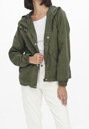 Femme portant une veste à capuche vert olive par-dessus un haut blanc et un pantalon blanc, avec la main gauche tenant la zone de la fermeture éclair de la veste.