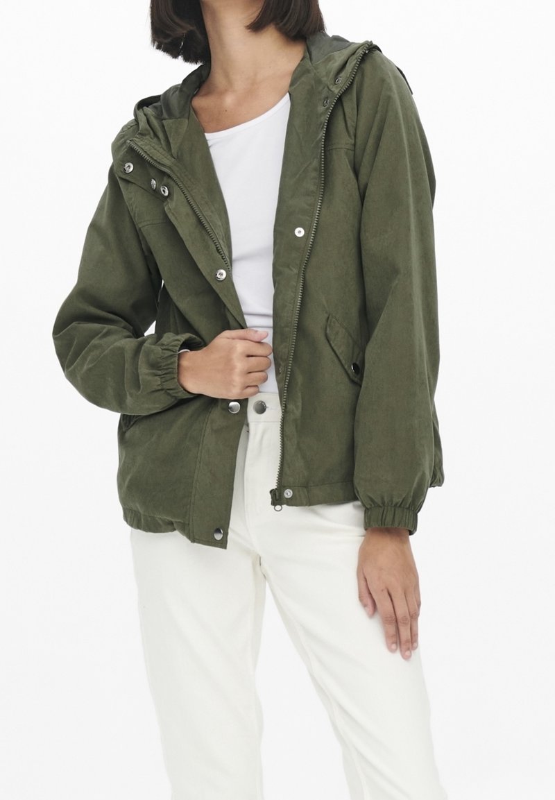 Femme portant une veste à capuche vert olive par-dessus un haut blanc et un pantalon blanc, avec la main gauche tenant la zone de la fermeture éclair de la veste.