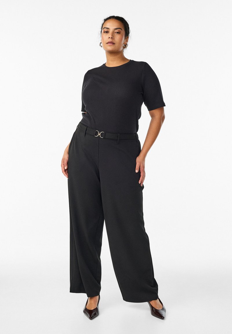 Kvinde står selvsikkert iført sort kortærmet top, sorte viddebenbukser med bælte, hoop-øreringe og sorte spidse højhælede sko på hvid baggrund.