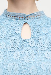 Κοντινό πλάνο ενός ατόμου που φοράει ένα ανοιχτό μπλε τοπ από δαντέλα με λουλούδια, με άνοιγμα στο λαιμό και λεπτομερή κεντημένο γιακά.