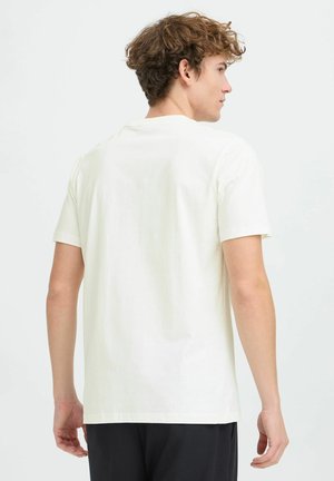 Young man with curly brown hair wearing a plain white short-sleeve t-shirt, facing away against a white background.