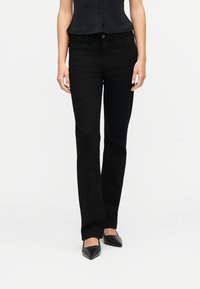 Γυναίκα που φοράει εφαρμοστά μαύρα τζιν με ίσια γραμμή, μαύρο αμάνικο τοπ και μυτερά μαύρα παπούτσια, στέκεται μπροστά σε ένα απλό λευκό φόντο.