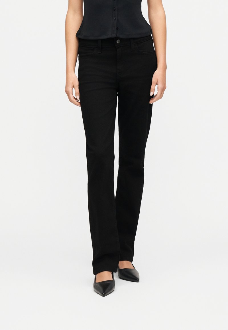 Γυναίκα που φοράει εφαρμοστά μαύρα τζιν με ίσια γραμμή, μαύρο αμάνικο τοπ και μυτερά μαύρα παπούτσια, στέκεται μπροστά σε ένα απλό λευκό φόντο.