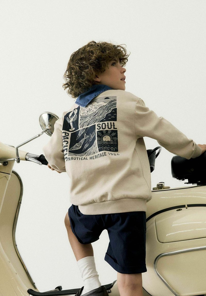 Sudadera beige con detalles en azul marino y un diseño gráfico en la parte de atrás, combinada con pantalones cortos azul marino y calcetines blancos. Scooter vintage al fondo.