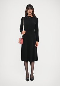 Abito lungo nero con maniche lunghe e colletto, caratterizzato da dettagli di bottoni sottili. Abbinato a una piccola borsa rosa e tacchi neri.