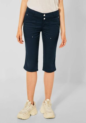 Frau trägt dunkelblaue Shorts bis zum Knie und beige klobige Turnschuhe, steht vor einem schlichten hellen Hintergrund.