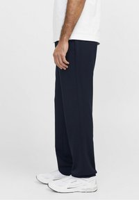 Uomo che indossa pantaloni blu navy, sneakers bianche e una t-shirt bianca, in piedi di lato contro uno sfondo chiaro e uniforme.