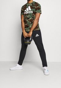 Camo-patroon groene Adidas T-shirt met wit logo, gecombineerd met zwarte sweatpants met wit logo en zwarte sneakers. Houdt een kettlebell vast.