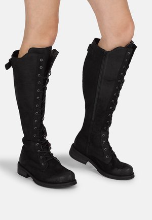 Botas de ante negras hasta la rodilla con cordones en la parte delantera y cremalleras laterales, con una suela plana y un diseño de puntera redonda.