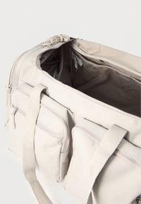 Μπεζ τσάντα τύπου duffel από καμβά με δύο εμπρόσθιες τσέπες με φερμουάρ, ανοιχτό κύριο διαμέρισμα και μπεζ υφασμάτινες λαβές σε λευκό φόντο.