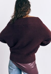 Kvinde med krøllet hår iført en løs mørk bordeaux off-shoulder sweater over en bordeaux lædernederdel og lyseblå jeans, set bagfra.