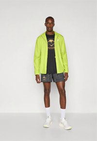 Lightweight, neon green jacket over a black graphic t-shirt and patterned shorts. White socks and athletic shoes complete the outfit.