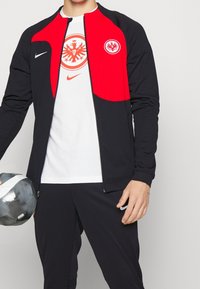 Chaqueta de chándal Nike en negro y rojo con cierre de cremallera, que presenta un logo del equipo en el pecho. Acompañada de una camiseta blanca y un balón de fútbol.