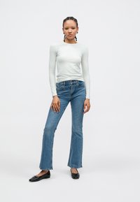 Dolga majica svetlo sive barve, oprijeta. Modre razširjene jeans hlače z nizkim pasom in posvetljenim vzorcem. Črne balerinke z mašnim detajlom. Preprost, klasičen slog.