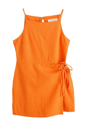 Kvinde i orange kjole og sorte sandaler står på stenetrin med en hvid og sort håndtaske, omgivet af hvide vægge og grønne planter.