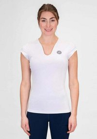 Mujer vestida con una camiseta blanca ajustada de cuello en V con un pequeño logotipo redondo en el lado izquierdo del pecho y pantalones azul oscuro, sonriendo a la cámara.