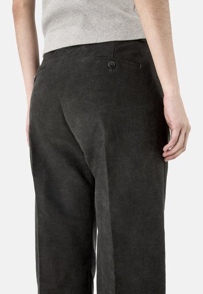 Osoba ubrana w czarne spodnie z zapinanymi na guziki tylnymi kieszeniami oraz jasnoszary top, widziana od pasa do połowy uda od tyłu.