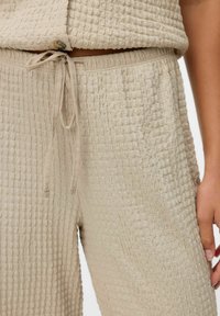 Beige textuur loungewear set met een top met knoopsluiting en losse broek met een elastische tailleband, beide in een gekreukt stof.