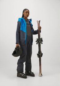 Ski gear featuring a navy and blue jacket, gray outfit, black ski boots, and helmet; holding skis with bindings, standing on a neutral background.