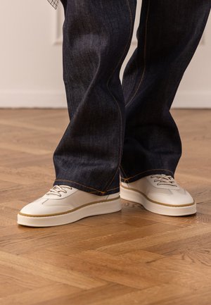 Personne portant un jean bleu foncé et des baskets en cuir blanc cassé avec des semelles beiges, debout sur un sol en bois.