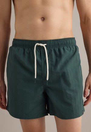 Torso masculino con pantalones cortos de baño verde oscuro con cinturilla elástica y cordón blanco, de pie frente a un fondo neutral.