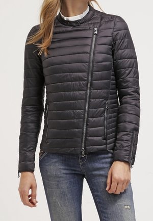 Frau trägt eine taillierte schwarze gesteppte Jacke mit asymmetrischem Frontreißverschluss und Reißverschlusstaschen an den Seiten, kombiniert mit blauen, zerrissenen Jeans.